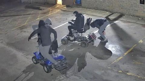 A night‑time CCTV image shows four people in a wet car park, all wearing hooded clothing and face coverings. They stand beside golf buggies.