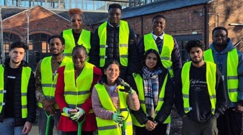 Ten people in hi-vis jackets are together standing up facing the camera. Buildings are behind them.
