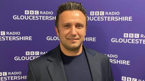 Dr Petse Alexovsky smiles looking at the camera, wearing a black jacket and black t-shirt in front of the BBC Gloucestershire back-drop