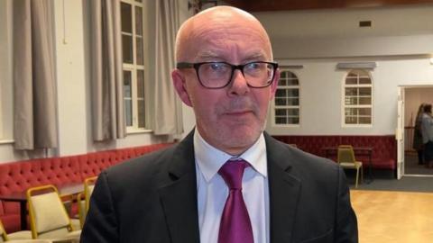 A headshot of a bald man who is wearing a black suit with a white shirt and cerise pink tie. He is smiling with his mouth closed and looking to the right of the shot. He is standing in a room with red cushioned benches around the edge and a light wooden floor in the centre. He is also wearing black rectangular glasses. 