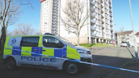 A police van sits behind blue and white police tape which is cordoning off a block of flats