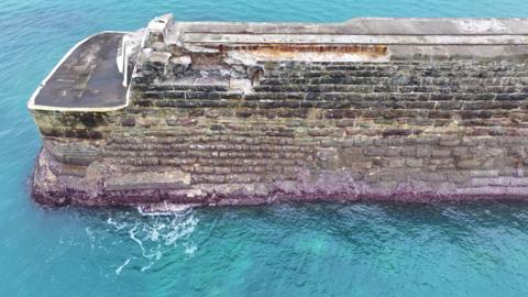 A breakwater of grey bricks is seen surrounded by blue-green sea, with damage to the breakwater visible in the form of missing sections. 