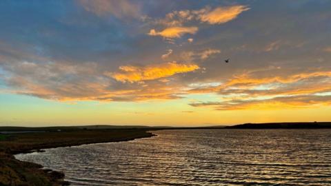 The sky is filled with yellow and grey clouds as the sun sets over the sea