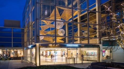 The entrance of Centre:MK at dusk. The entrance is glass with several doors, large round lights inside with a tree to the right with lights in it. It is a square building and several people are visible inside.