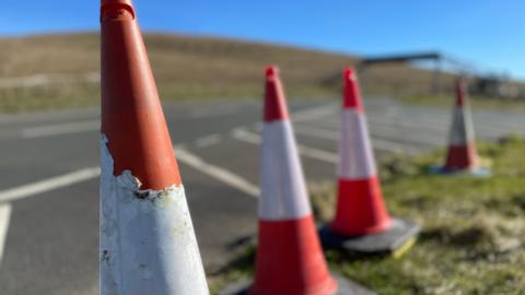 A close up of orange and white cones along the roadside, you can see a metal bridge in the background to cross the road.