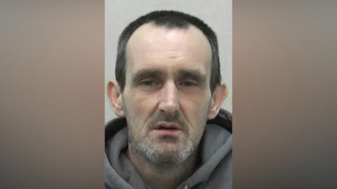Mugshot of Young. He is a dishevelled looking man with pale pasty skin, a glazed expression on his eyes. He has grey stubble and short black hair.