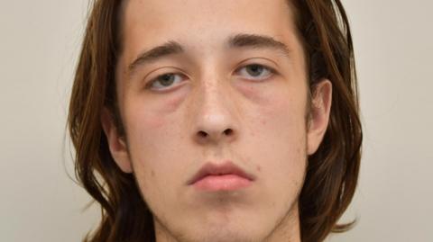 Alfie Coleman has shoulder-length brown hair swept backwards. He looks tired but shows no emotion while pictured in this custody mugshot.