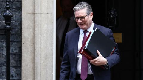 keir Starmer pictured leaving Number 10,  wearing a navy suit with purple tie, carrying folders under his arm. He is looking  down at the ground.