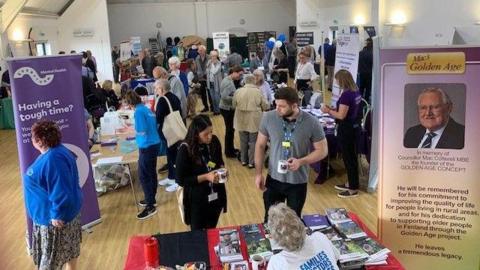 People have gathered in a hall, there are tables with leaflets on them and banners.