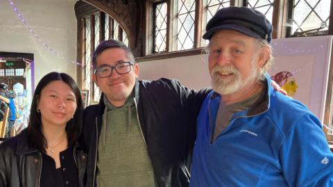 Suzanne Chung, Ed Ngai and Chris Turland stand smiling, with book shelves and art work behind them.