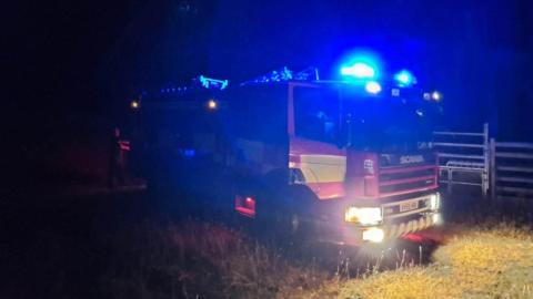 A fire engine at night parked up in a field. The vehicle's blue lights are flashing and it is only illuminated by its headlights. It is predominately red with yellow high viz squares down its side. 