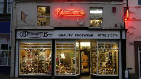 A shop with wide windows that display types of shows and has a white sign which says BRASSINGTON BROS - QUALTITY FOOTWEAR and has the number 01782215570 all in dark blue lettering. There is a bright red Brassington sign above the door.