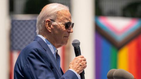 Joe Biden holds a microphone while speaking in front of a Pride flag