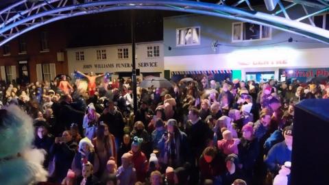 A night-time view across a street from a stage, showing the back of Father Christmas's head to the edge of the foreground and hundreds of people facing the stage. A row of shops stands behind the crowd