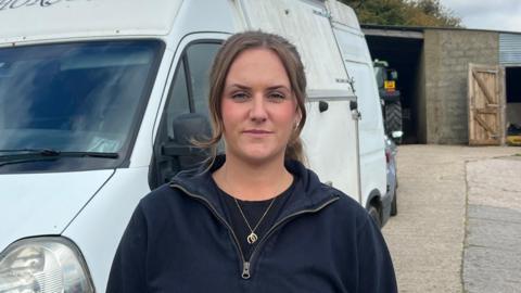 Ceylone Fletcher stands in front of a white horsebox van, with part of the word 'Horses' visible on the top. She is wearing a dark sweatshirt and stood in a rural setting, with a stable behind her and a part of a tractor visible. The sky is partly cloudy, and there are trees in the distance.