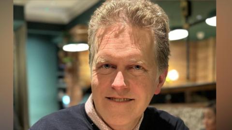 Charles Allen smiles at the camera while sitting in a restaurant. He is holding a small glass with bright red liquid inside it along with large ice cubes. He has light blonde hair with some grey in it. He wears a navy blue jumper with a pink shirt underneath.