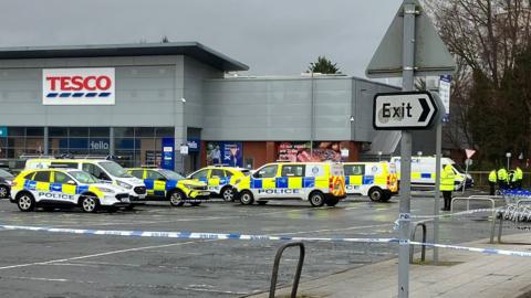 Police vehicles cordon off a Tesco supermarket car park with blue‑and‑white tape as officers stand nearby under a grey sky.