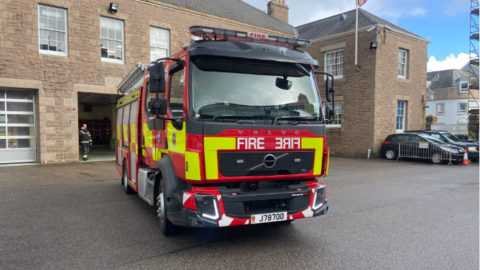 A large fire vehicle parked in front of a large brown building - the fire station. Normal cars can be seen parked to the right.