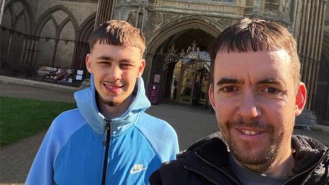 Kevin Lacey is in a dark coat and has short hair. He is pictured standing next to his son Ethan who is in a blue tracksuit top with a moustache and small beard. 