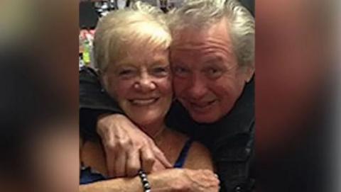 Sheila Jackson with blond hair smiles as Eric Greener with grey hair has his arm around her neck in front of a bar. Eric is also smiling.