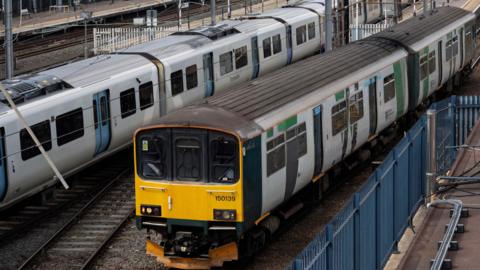 Two trains in Bedford railway station, showing one for the Marston Vale line. There are power cables, tracks a yellow train carriage. Metal fences, to the right and platforms to the right.