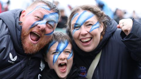 Bath fans - a man, a boy and a woman - wearing blue and white face paint with mouths open