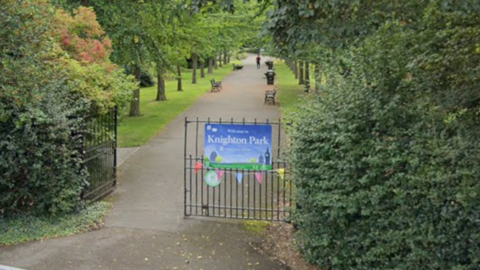 The entrance to a park, with black metal gates and a park sign stuck to the gates, leading to a pathway lined with benches and mature trees