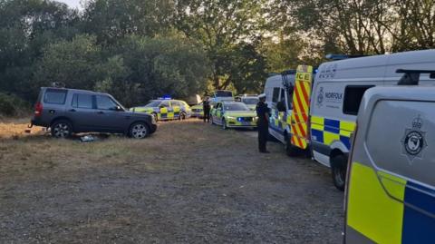 Seven police vehicles are parked on a road, blocking it off entirely. They are a mix of police cars and riot vans. There are two officers, dressed in black, standing next to the vehicles. To the left, further on the grass, there is a dark grey off-road vehicle.