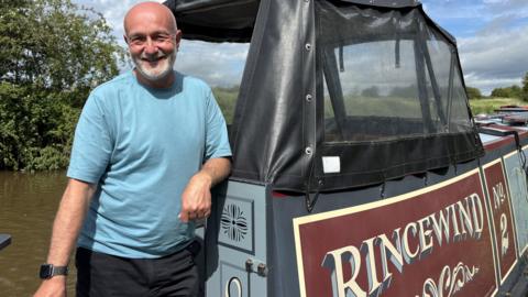 A bald man with white facial hair is smiling and standing on a canal boat. He is wearing black shorts and a light blue t-shirt. His boat is blue grey in colour and on the side of it is its name, painted in yellow writing on a wine red background. It reads "Rincewind number two"