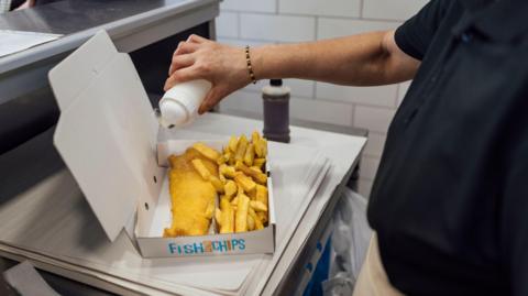 A view over a person's shoulder of a cardboard box of fish and chips on paper and a stainless steel counter, with a hand sprinkling salt over the food. A bottle of vinegar can be seen in background.