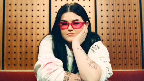 Mae Stephens has long dark hair, pink glasses, and sits in a recording studio, leaning forward with one arm resting on a table and the other propping up her face. She is wearing a light-coloured sweatshirt covered in handwritten-style illustrations and text, with several visible tattoos along her arms. Behind her is a wooden acoustic panel wall with evenly-spaced circular sound‑dampening holes, giving the space a warm, studio-like atmosphere.