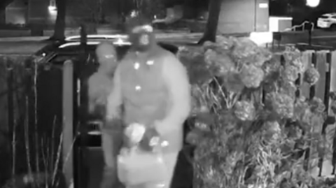 Black and white CCTV footage of two men in balaclavas holding what appears to be petrol cans walking through a front garden gate.