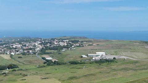 A picture taken from above looking down across Alderney. It is an island with a lot of grassland and houses in the distance. 