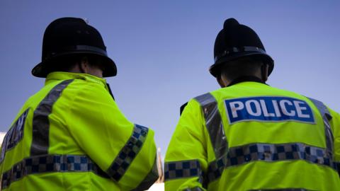 The back of two police officers wearing police hats and a yellow police jacket. A blue sky is in front of them.