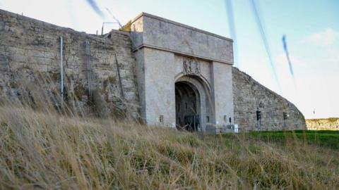 A general view picture of HMP The Verne in Portland, taken on 4 February 2022. The picture shows the entrance to a stone tunnel, with a coat of arms above it, and a stone wall on either side of it.