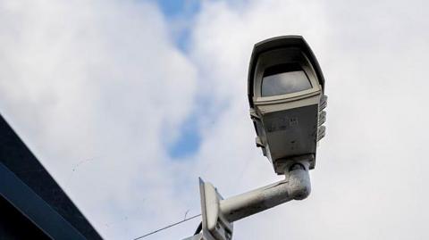 A CCTV camera on the side of a building. The sky is blue with clouds.