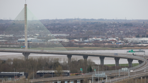 The Mersey Gateway toll bridge, which runs across the River Mersey. It is a cable-stayed bridge with the access road looping round from the west.  