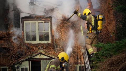A firefighter stands on a ladder as they move burned straw from a thatched roof that is on fire. Smoke can be seen billowing from the property. Another firefighter is working on the ground underneath the one on the ladder.