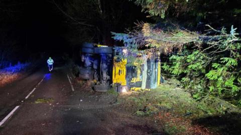 A gritter lorry on its side on a roadside in the early hours. It is dark and there is lighting on it.