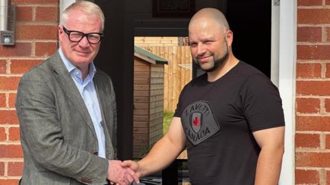 A man in a grey suit jacket shaking hands with a man in a black t-shirt, both smiling, outside the front door of a house.