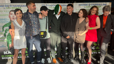 The cast and director of the Kneecap film standing in a line in front of posters for the movie