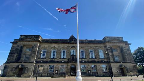 A wide shot of Wirral Council's building. It is grand but the brick work is weathered. There is a flag pole out on the front of the site flying the Union flag.