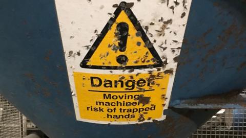 A worn-out warning sign on a piece of machinery that has a black exclamation mark in a yellow triangle with a black edge. It reads Danger. Moving machinery - risk of trapped hands