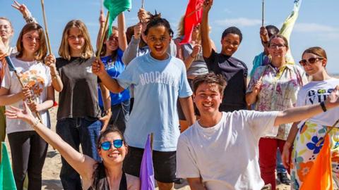 A group of young people on a sandy beach at Great Yarmouth.  They are wearing summery clothes and are waving colourful flags and looking happy. The sky behind them is blue.