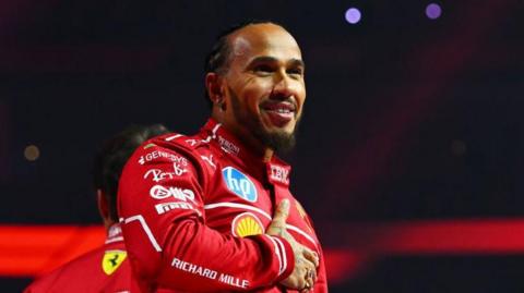 Lewis Hamilton holds his right hand across his chest while smiling at the F1 2025 season launch event. He is wearing the red Ferrari uniform