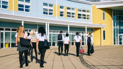 Pupils in school uniform walk towards a school building in the sun. 