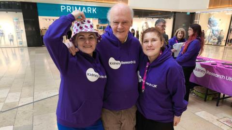 Three volunteer (two women and one man) wearing purple jumpers with "KidneyResearchUK" written on them smile at the camera. They are standing in the atrium of the shopping centre. 