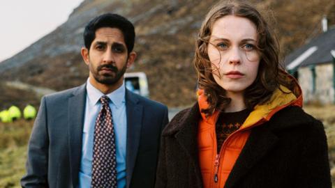 The Islands' DCI Ahmed Halim played by Sagar Radia and dectective Kat Crichton played by Sorcha Groundsell stand together in the outdoors. Behind them is a cottage, a police vehicle and a rugged hillside.