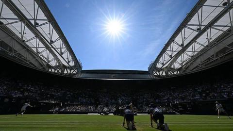 The court can be seen with players in action and with the roof open. There is a bright blue sky and bright sun 