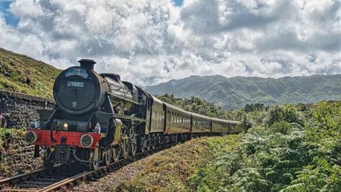 A steam train called The Jacobite comes towards us through a lush Highland landscape.